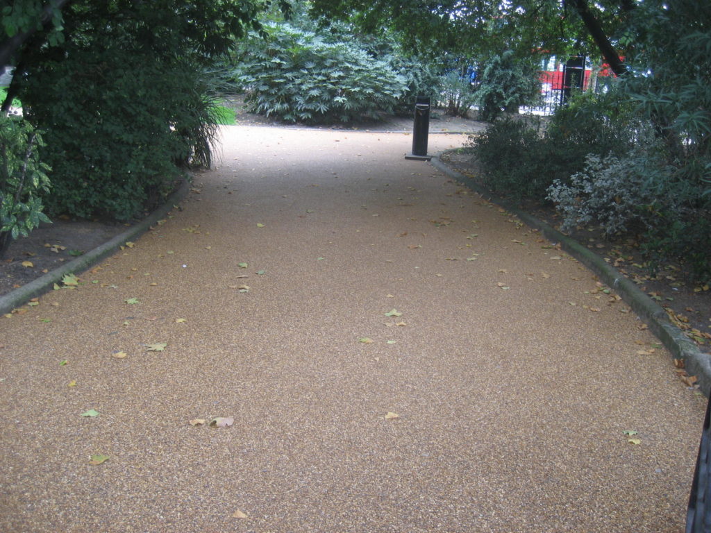 A wide, empty pathway made of light brown resin-bound gravel extends through a green park or garden, with trees and bushes lining both sides of the path.