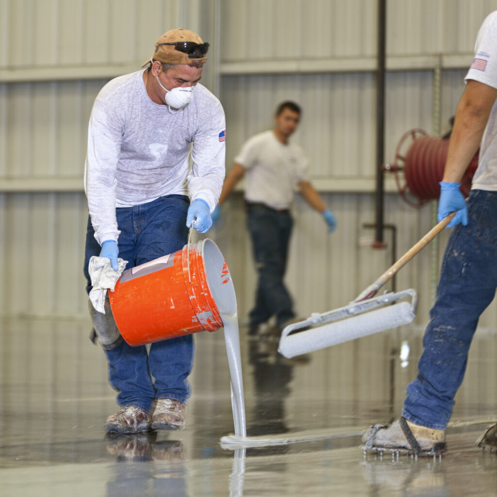a man pours epoxy on a floor while others wait to roll it on the floor at a warehouse