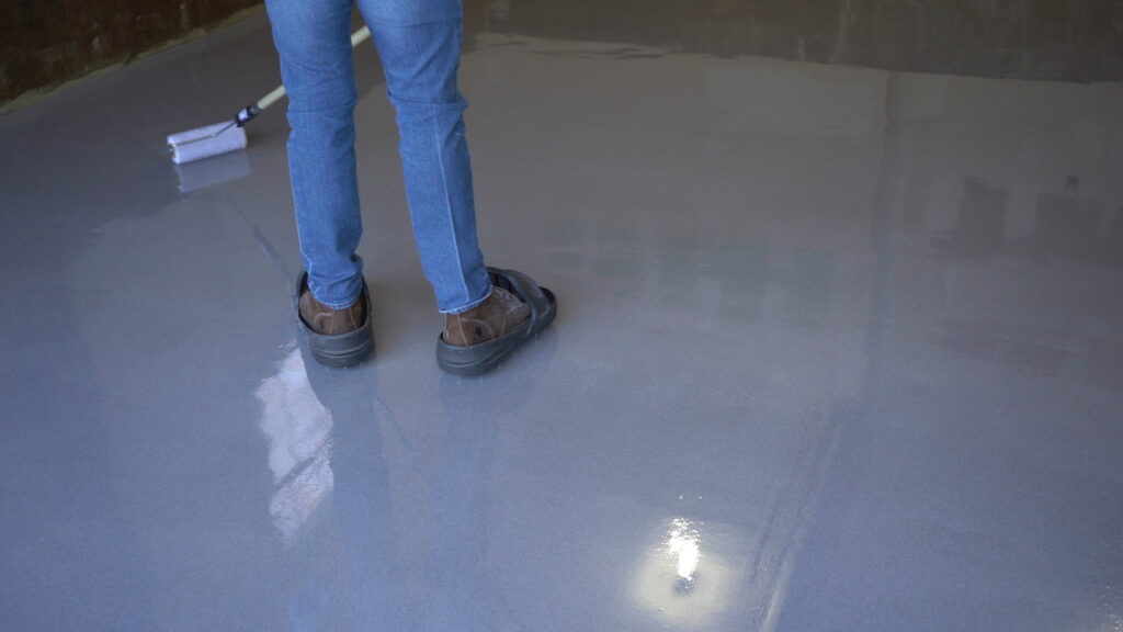 A low-angle close-up shot of a person standing on a glossy gray epoxy floor. The person is holding a long-handled paint roller and has just rolled a new section of the floor, which is visible in the upper-left corner of the frame.
