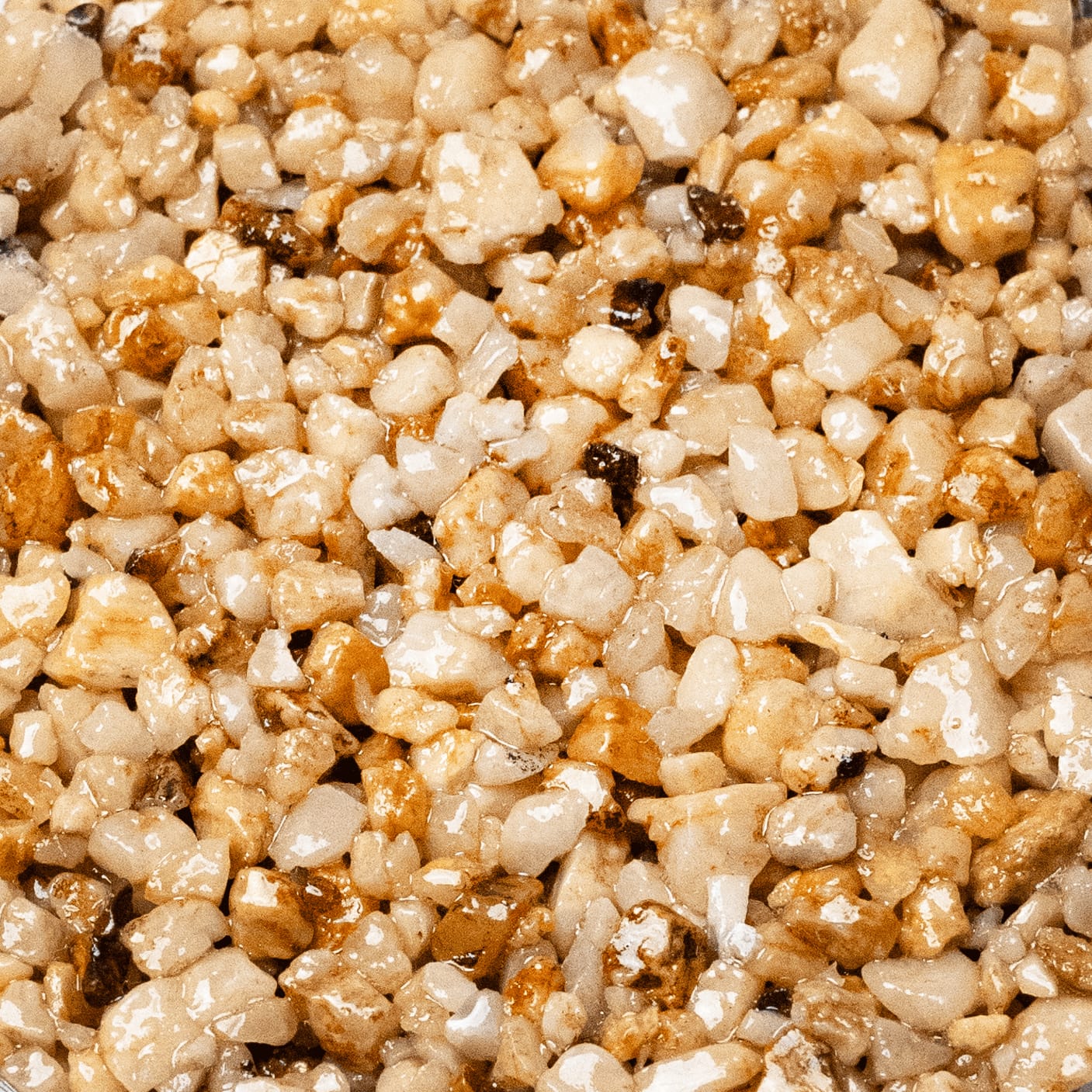 Aut Quartz 1. A close-up view of wet, beige and amber-toned gravel. The small, angular stones have a glossy, reflective surface, suggesting they are covered in a clear sealant. The colors are predominantly light, with a mix of creamy white, beige, light brown, and some darker, amber-toned pieces.