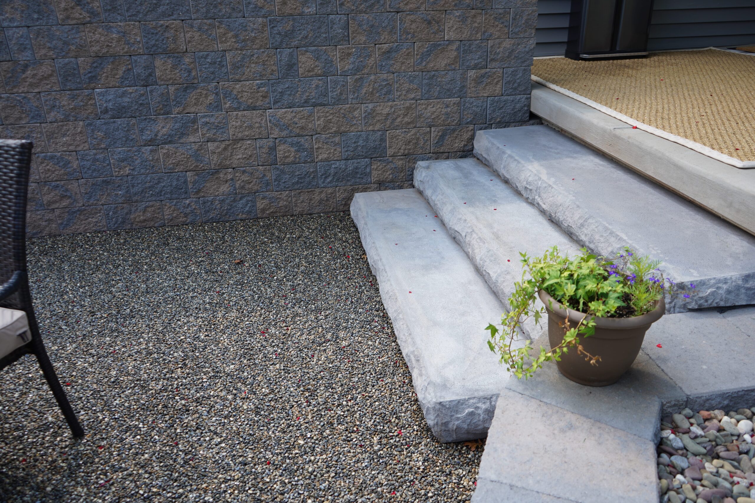 A view of an outdoor patio featuring a set of three light grey concrete steps leading up to a door. The steps have a natural, textured, and slightly rough finish. To the left of the steps, the ground is covered with a dark grey and tan gravel-like surface.