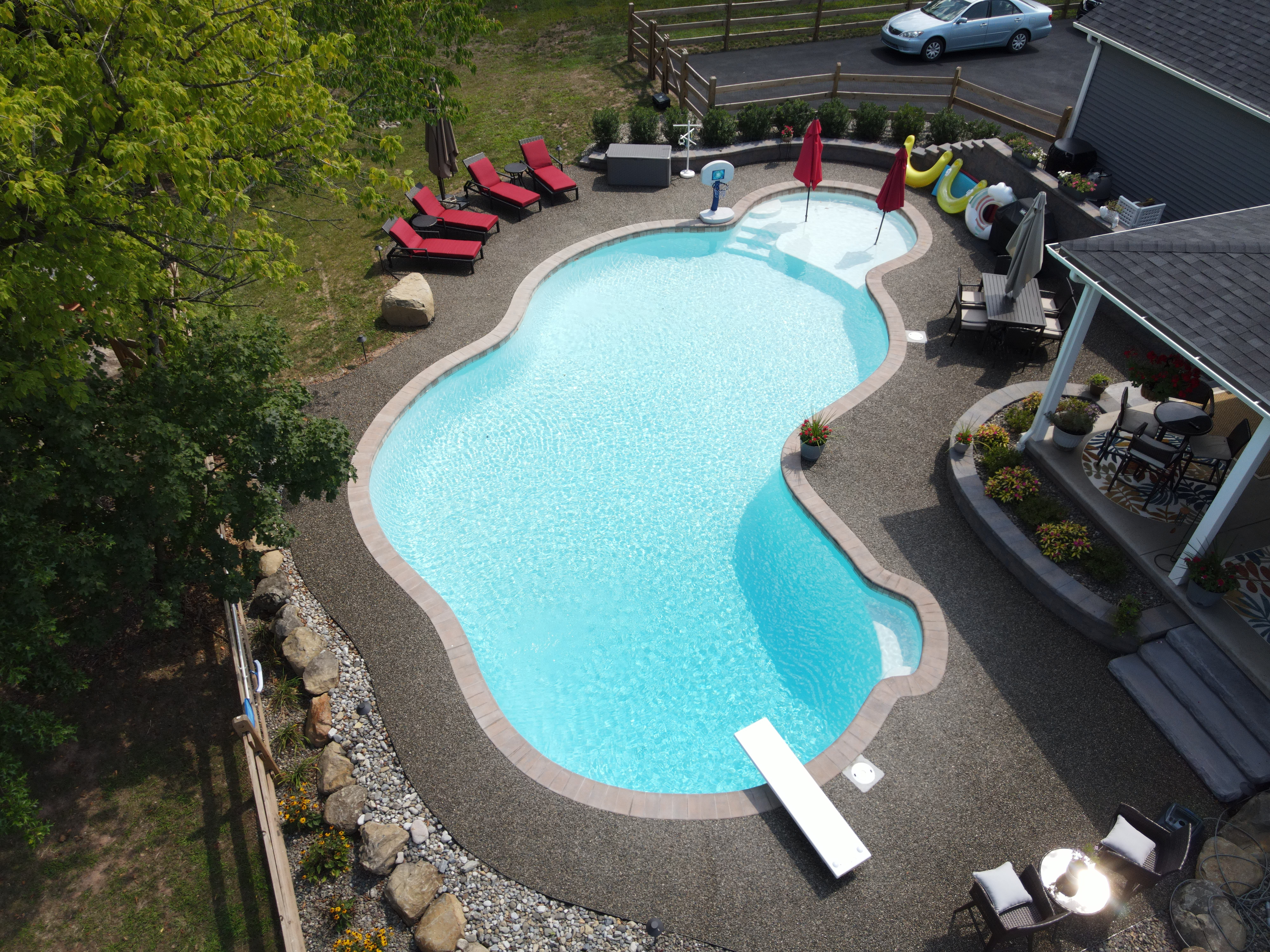 An aerial view of a large, kidney-shaped backyard swimming pool with a light blue interior. The pool is surrounded by a textured, dark grey and brown gravel deck.