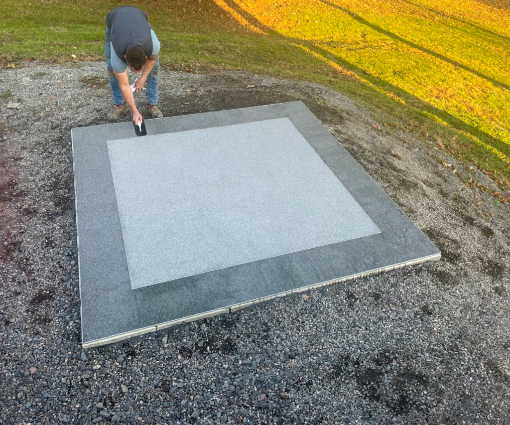A high-angle shot shows a man on a gravel-covered ground using a tool to smooth a gray surface. The surface is a large, square slab, with a lighter-colored square inset in its center, creating a two-toned pattern.