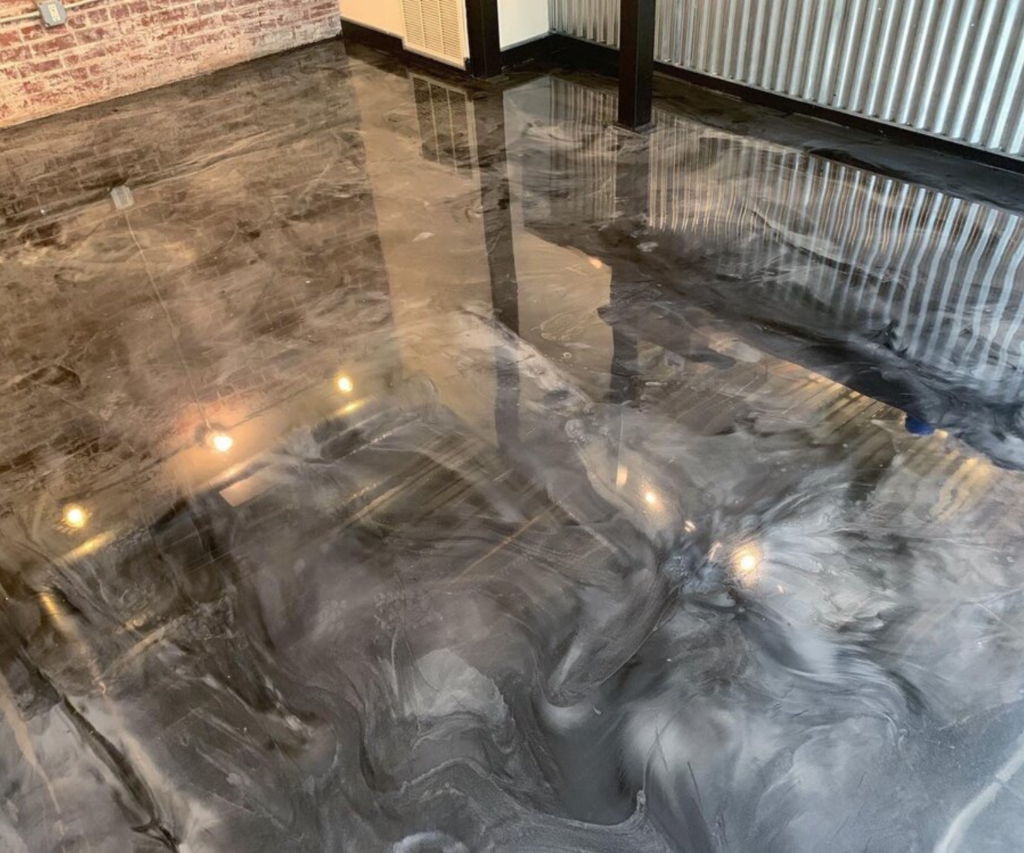 A glossy, dark metallic epoxy floor with a swirling pattern of black, gray, and white. The surface is highly reflective.