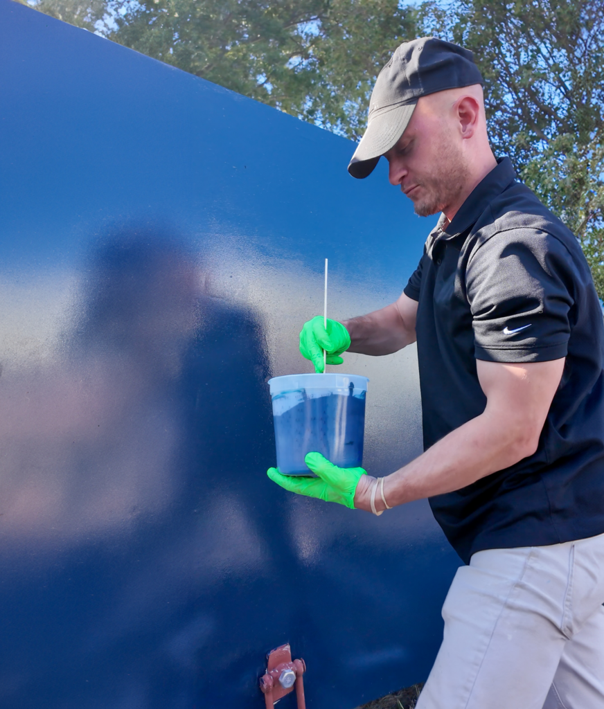A man holding a clear plastic bucket containing a thick, dark blue substance. He is stirring the mixture with a white stick and standing next to a large, glossy, dark blue wall.