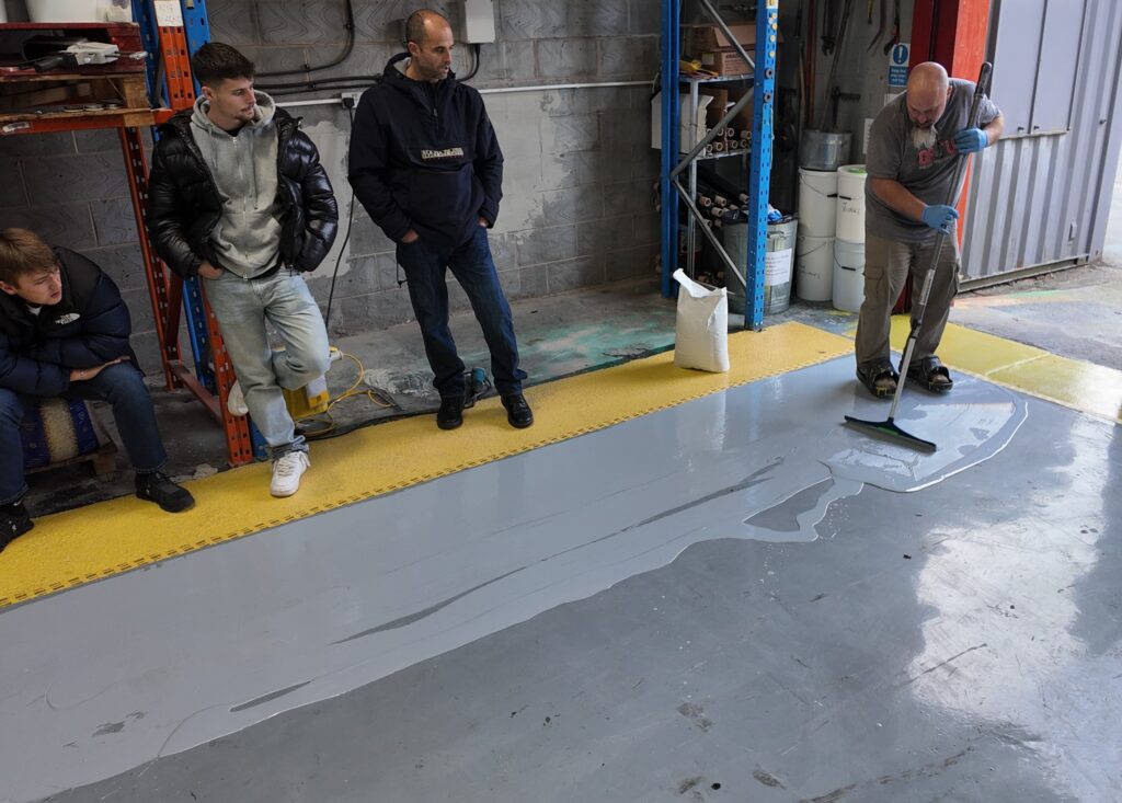 A group of people in a warehouse setting, observing a man applying a gray liquid coating to the floor using a long-handled squeegee. The liquid is spreading across the concrete floor, leaving a glossy finish.
