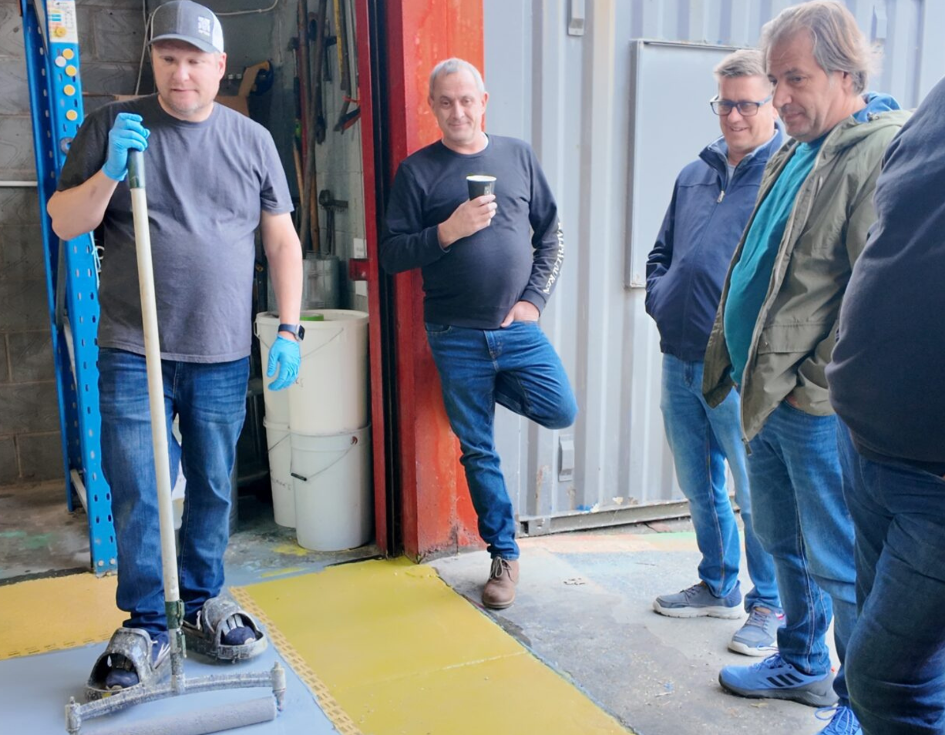 A group of five men standing in a workshop, with one man in the foreground holding a long-handled floor roller. The floor is being resurfaced with a gray coating, next to a yellow, tiled section. The other men are observing.