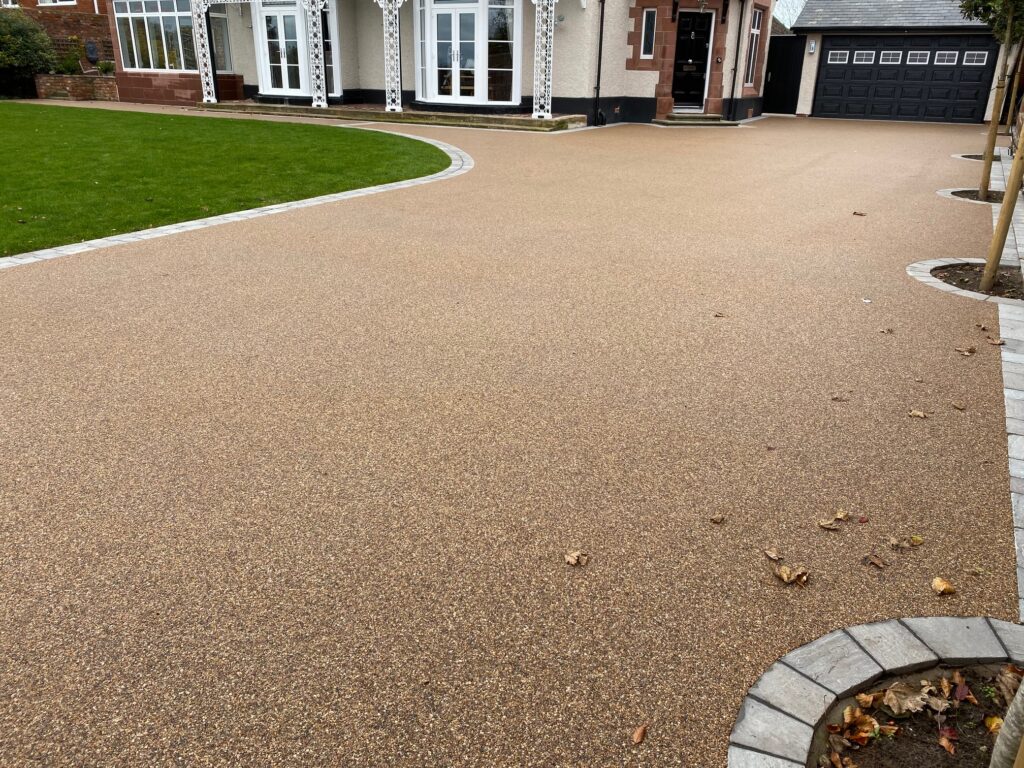 A large residential driveway and entrance area finished with a smooth, tan resin-bound gravel surface. The expansive driveway features a decorative grey stone border and leads to a white house with ornate porch pillars and a black garage door.