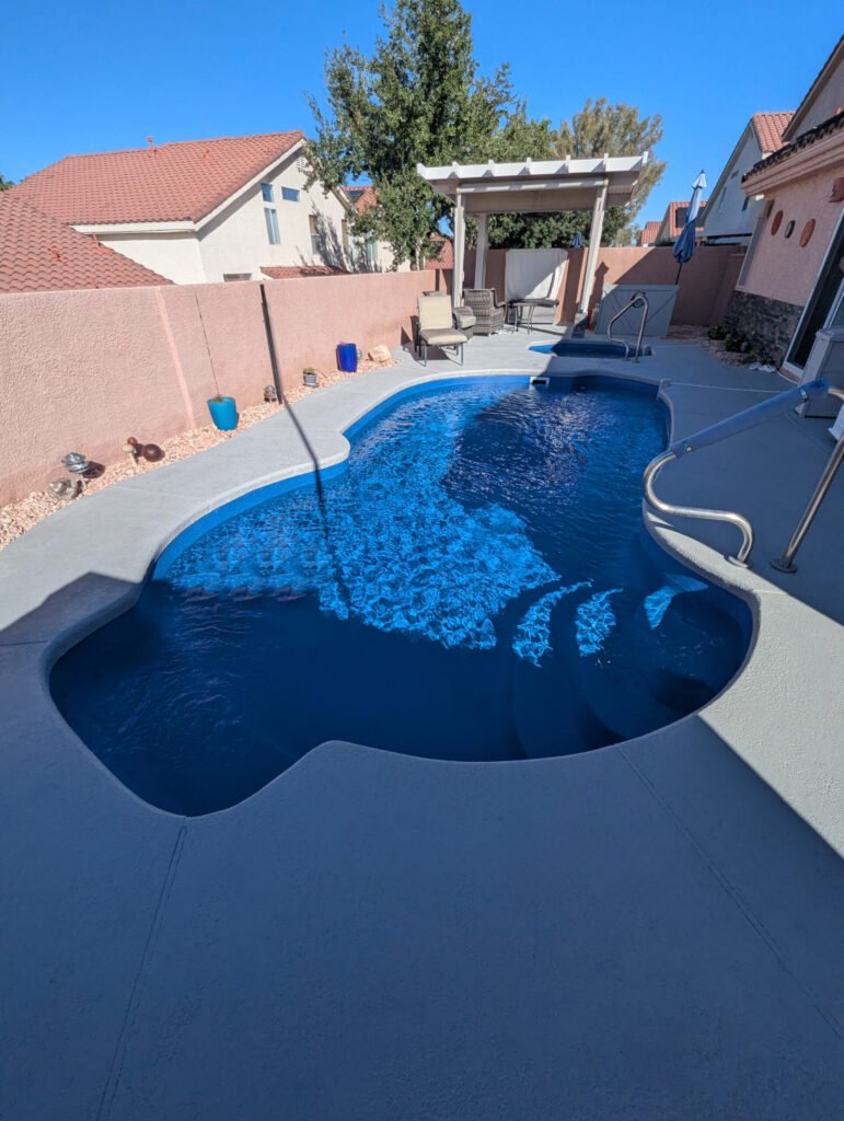 A high-angle shot of a curved, dark blue inground swimming pool with sparkling water. The pool is surrounded by a smooth, light grey concrete deck.