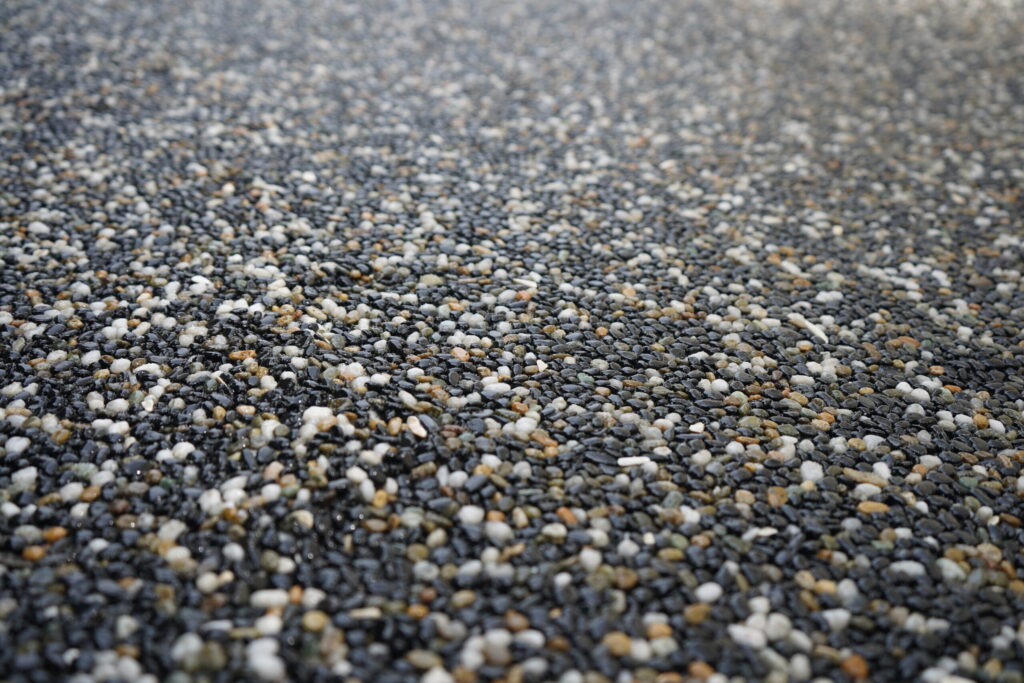 Close-up, shallow depth of field shot of a textured resin-bound aggregate surface. The surface is composed of small, polished stones in a mix of dark gray, black, white, and warm amber tones, tightly bound together with a glossy, wet-look finish. The foreground is in sharp focus, while the background softly blurs out.