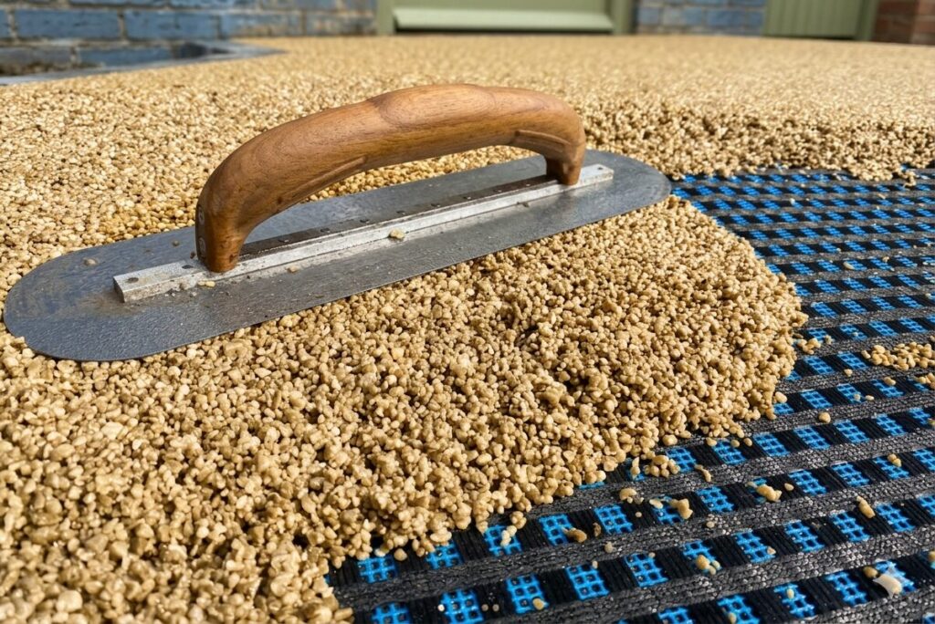 A close-up view of a hand trowel with a wooden handle resting on a pile of light brown resin-bound stone aggregate. The trowel is positioned at the edge of a black and blue reinforcement mesh grid, showing the process of spreading and compacting the mixture during a resin-bound surfacing installation.