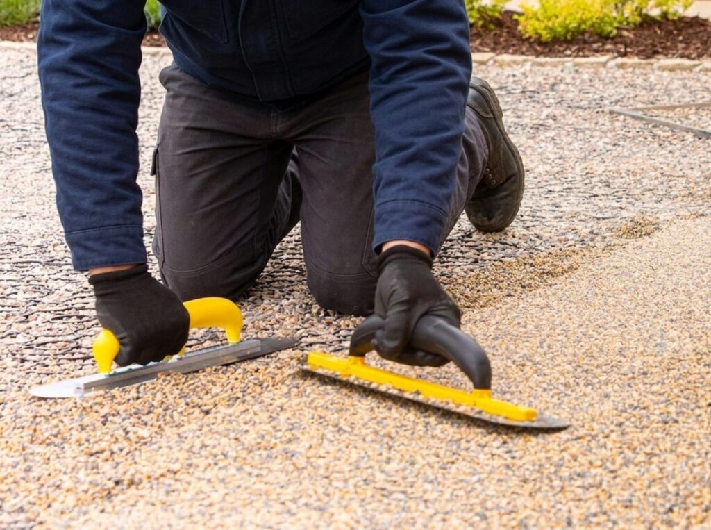 A person wearing a blue jacket, dark work pants, and black gloves is kneeling on a gravel surface while using two yellow-handled trowels to smooth or level the ground. One trowel is held in each hand as they work on a bed of small stones or aggregate.