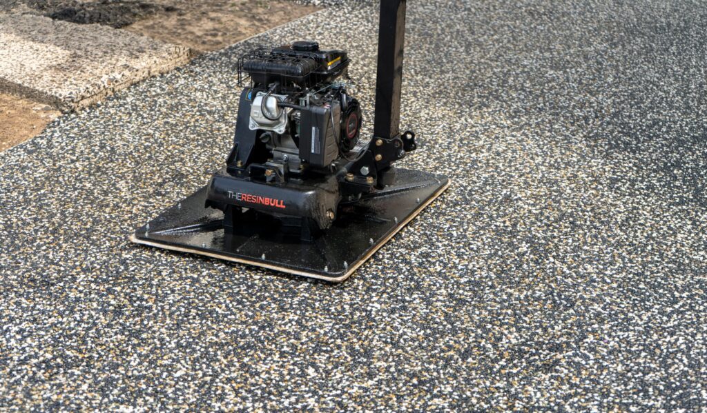 A black motorized plate compactor called Glidabull resting on a freshly laid resin-bound stone surface. The machine features a large flat base plate, a gasoline engine, and a tall handle, and is designed to compact and finish resin-bound gravel installations faster and more consistently than traditional hand troweling.